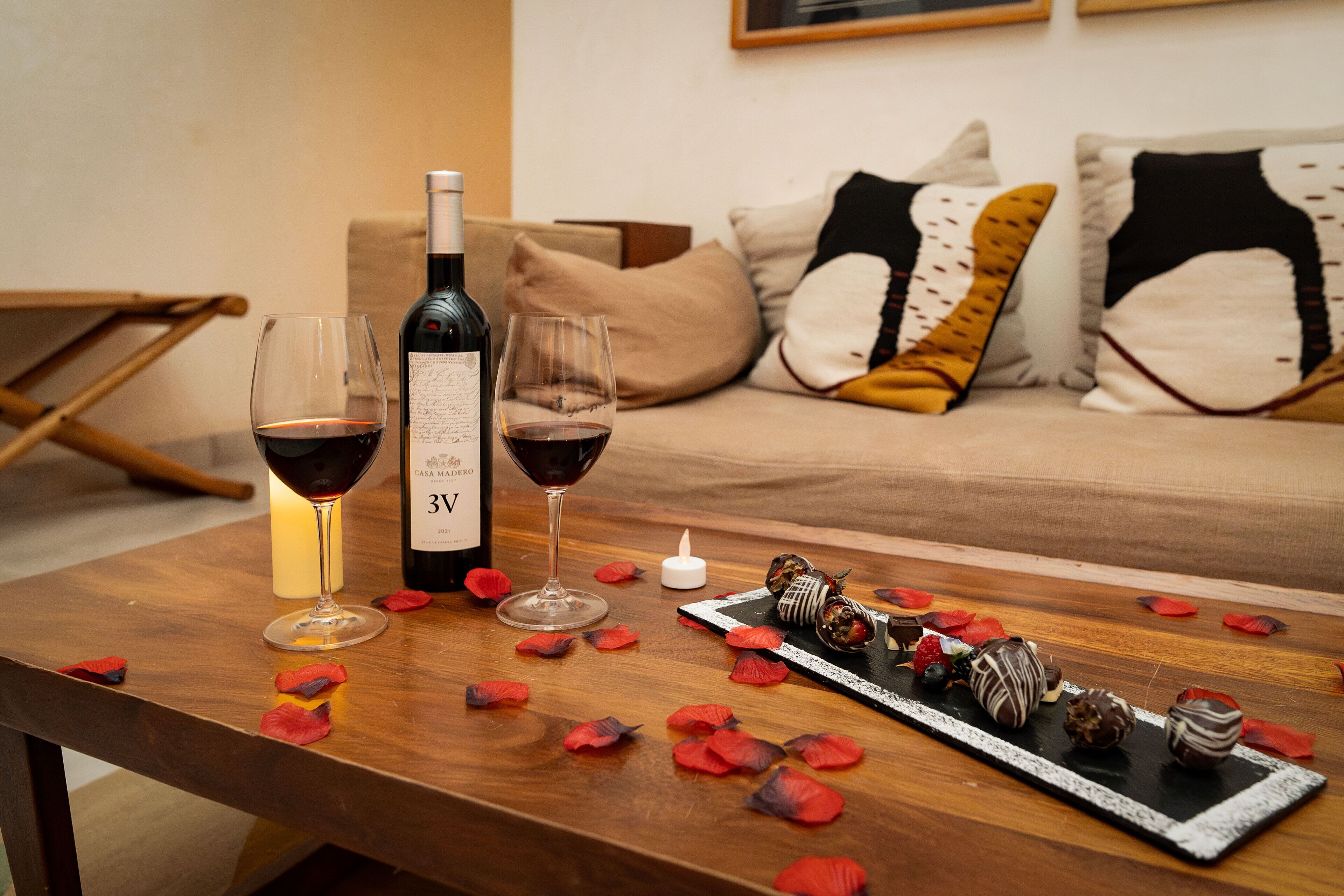 Een houten tafel met een fles wijn, twee wijnglazen, rozenblaadjes en chocolade.