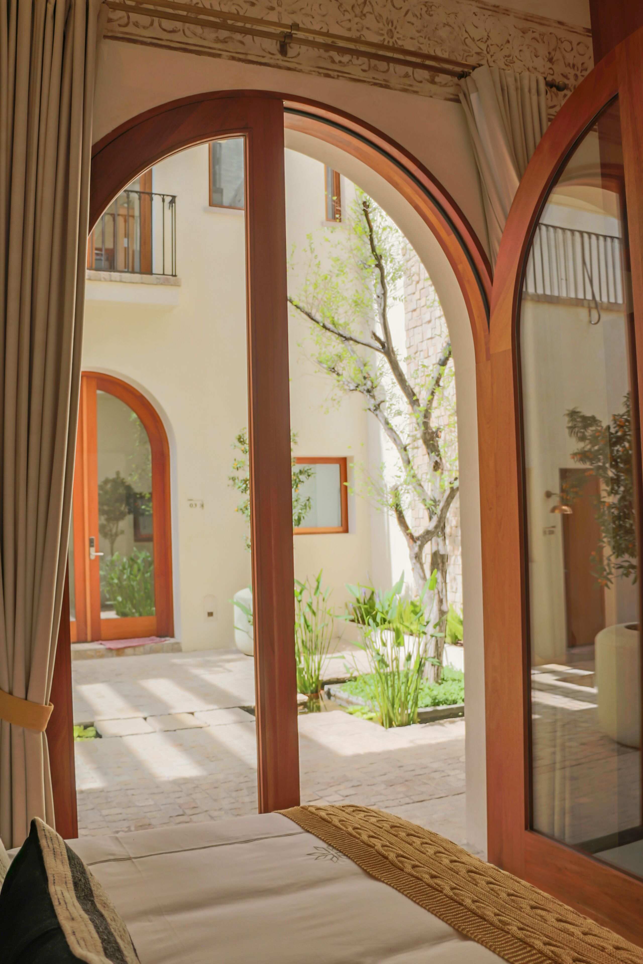 A room with a bed, a wooden door, and a view of a courtyard with plants and a building in the background.