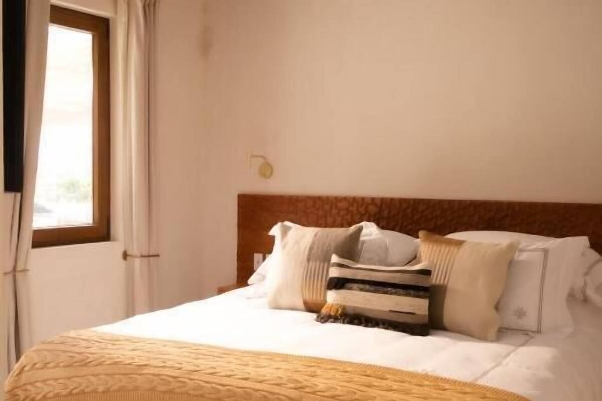 A bedroom with a wooden ceiling, a large bed with white bedding and a yellow throw, and a window with curtains.
