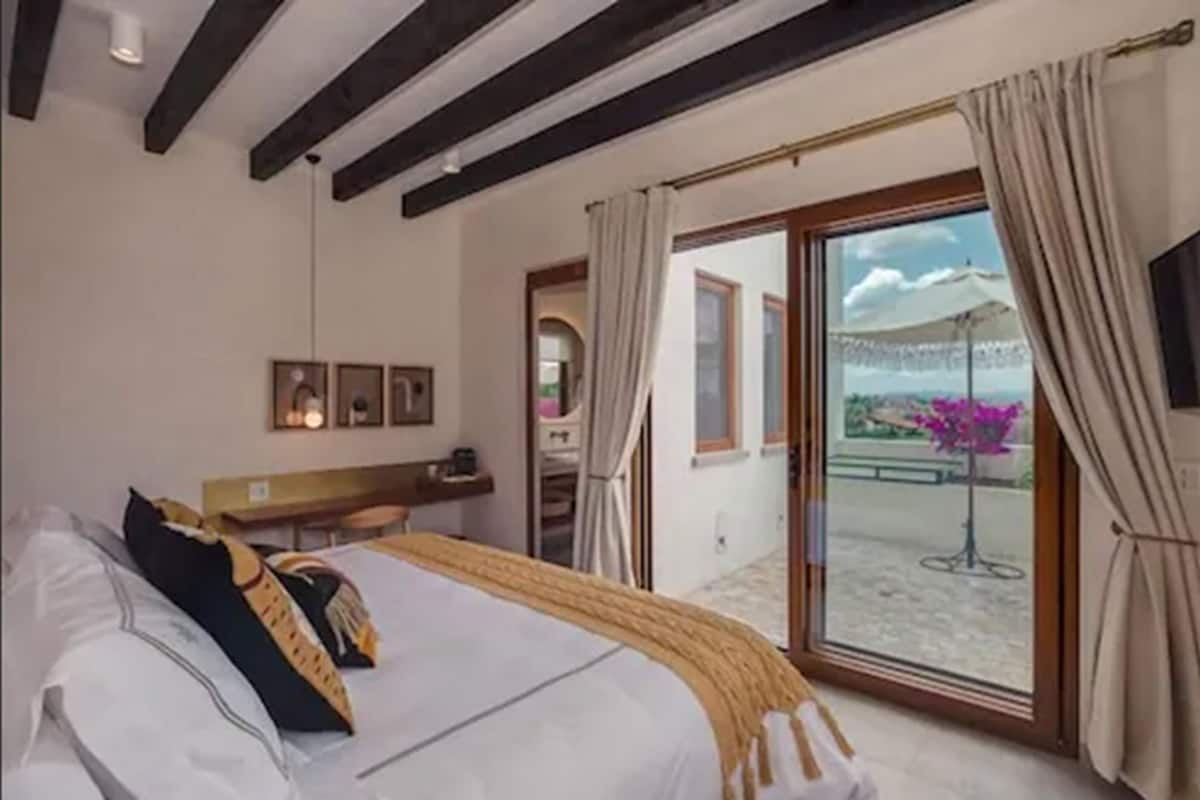A hotel room with a bed, a desk, a chair, a window with curtains, and a view of the outside.