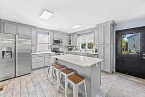 A modern kitchen with stainless steel appliances, a large island with bar stools, and a black door with a view of the outside.
