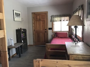 A small, cozy bedroom with a wooden door, a window with star-patterned curtains, a bedside table with a lamp, and a bed with a red blanket.