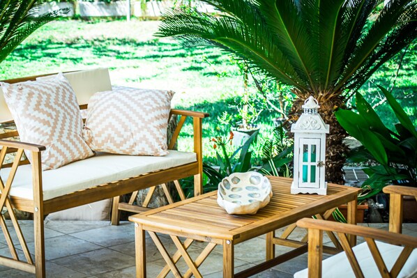An outdoor seating area with wooden furniture, including a sofa and a table, surrounded by lush greenery.