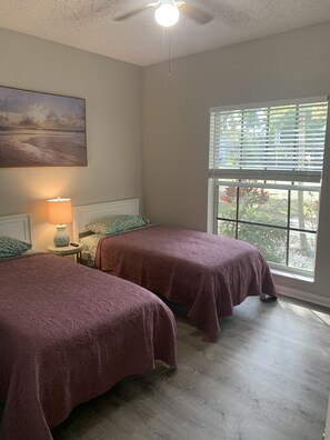 A bedroom with two beds, a ceiling fan, a window with blinds, and a painting on the wall.