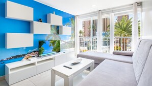 A modern living room with a white sofa, a coffee table, and a TV on a white stand. There are white shelves against a blue wall with a tropical mural. A balcony with a view of palm trees and buildings is visible through large windows. - Seimar 1-8 Apartment Levante Beach (Benidorm)
