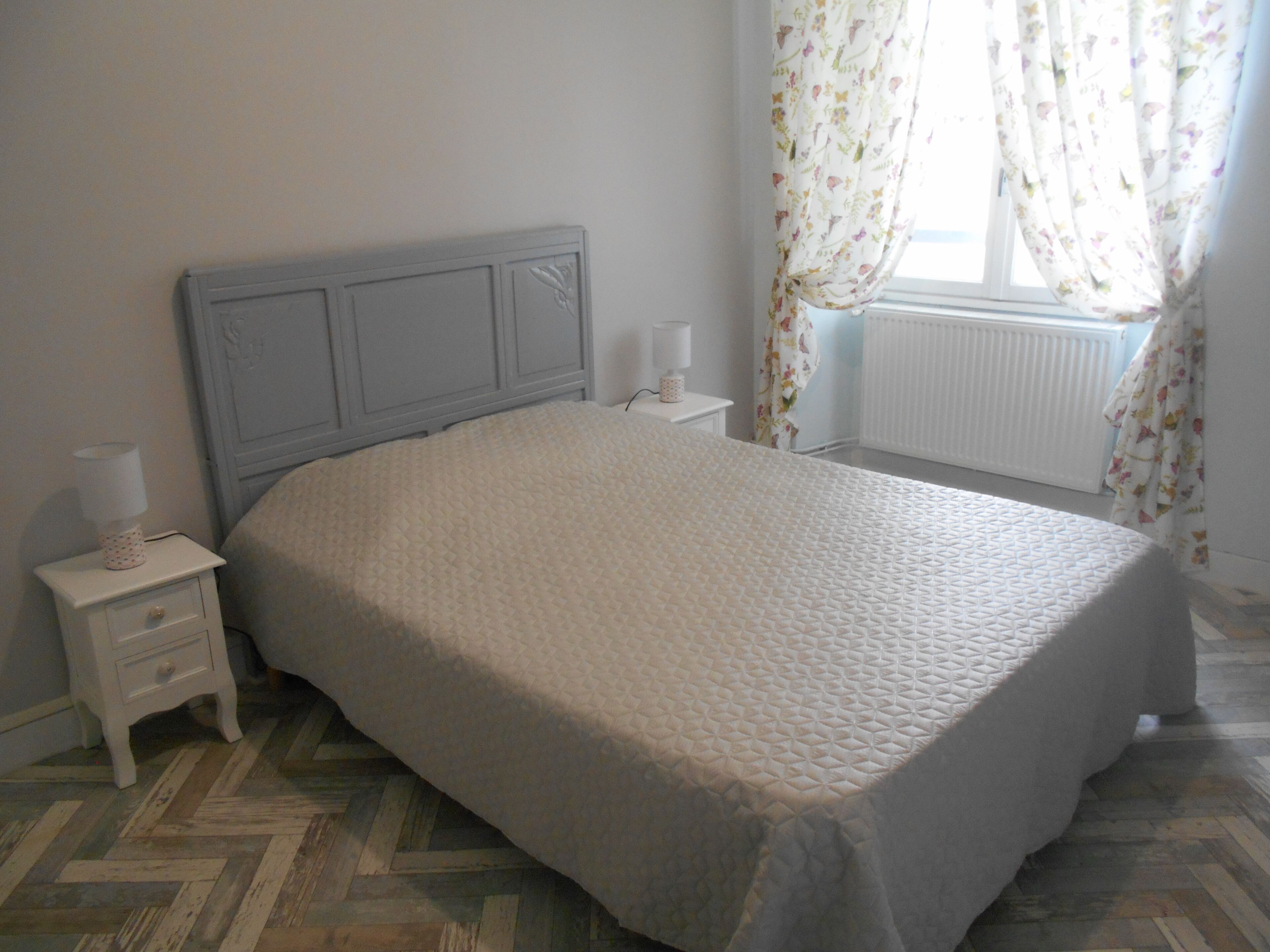 Une chambre à coucher avec un lit, une table de chevet, une lampe, un radiateur et des rideaux à motifs floraux.