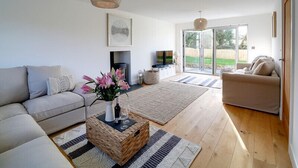 A modern living room with a sofa, a coffee table with a vase and wine glasses, a fireplace, and a large sliding glass door leading to a garden. - Elmwood (Port Isaac)
