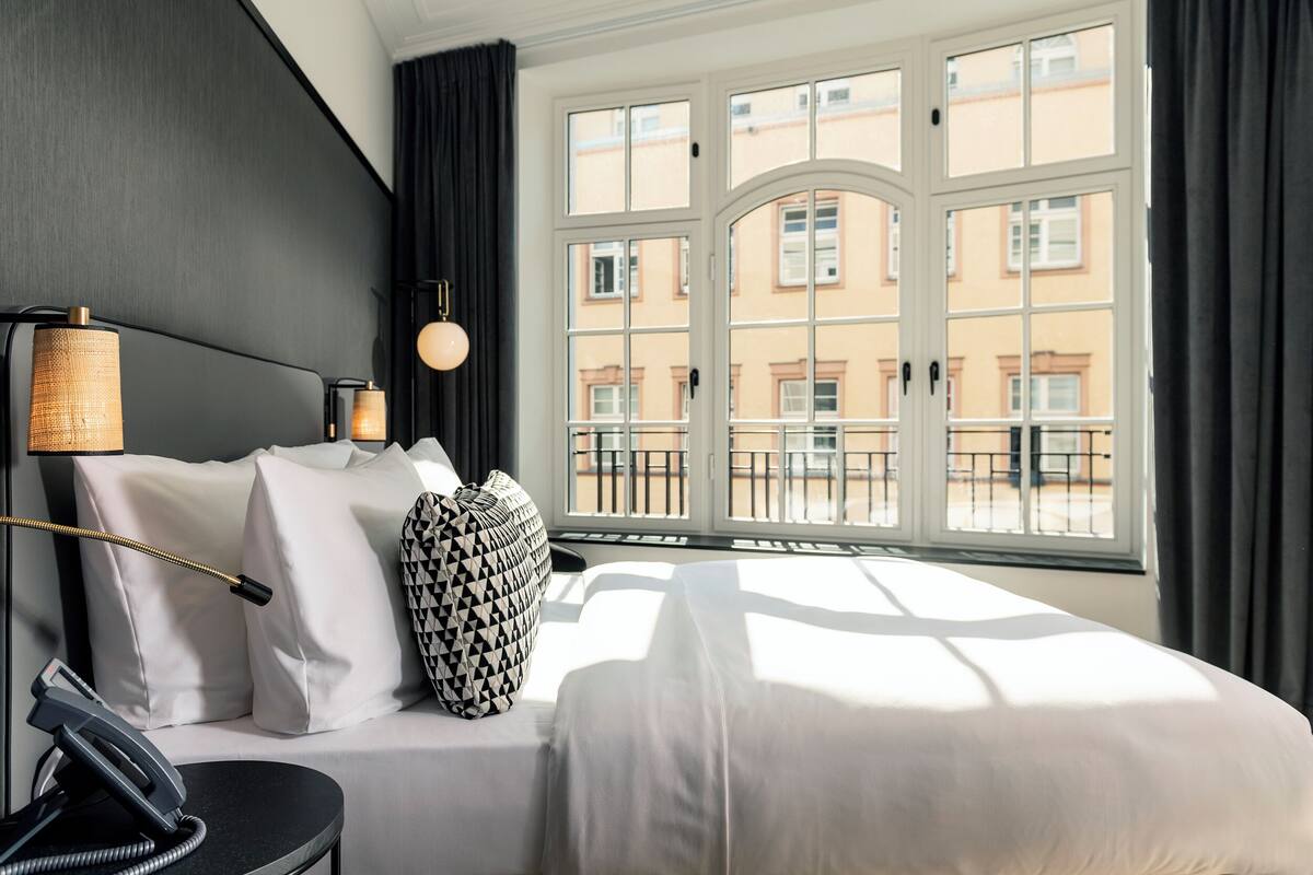 Quarto de hotel com uma janela grande, uma cama com roupa de cama branca, um telefone preto e uma almofada estampada.
