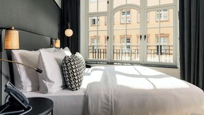 Habitación de hotel con un ventanal grande, cama con sábanas blancas, teléfono negro y almohada estampada.