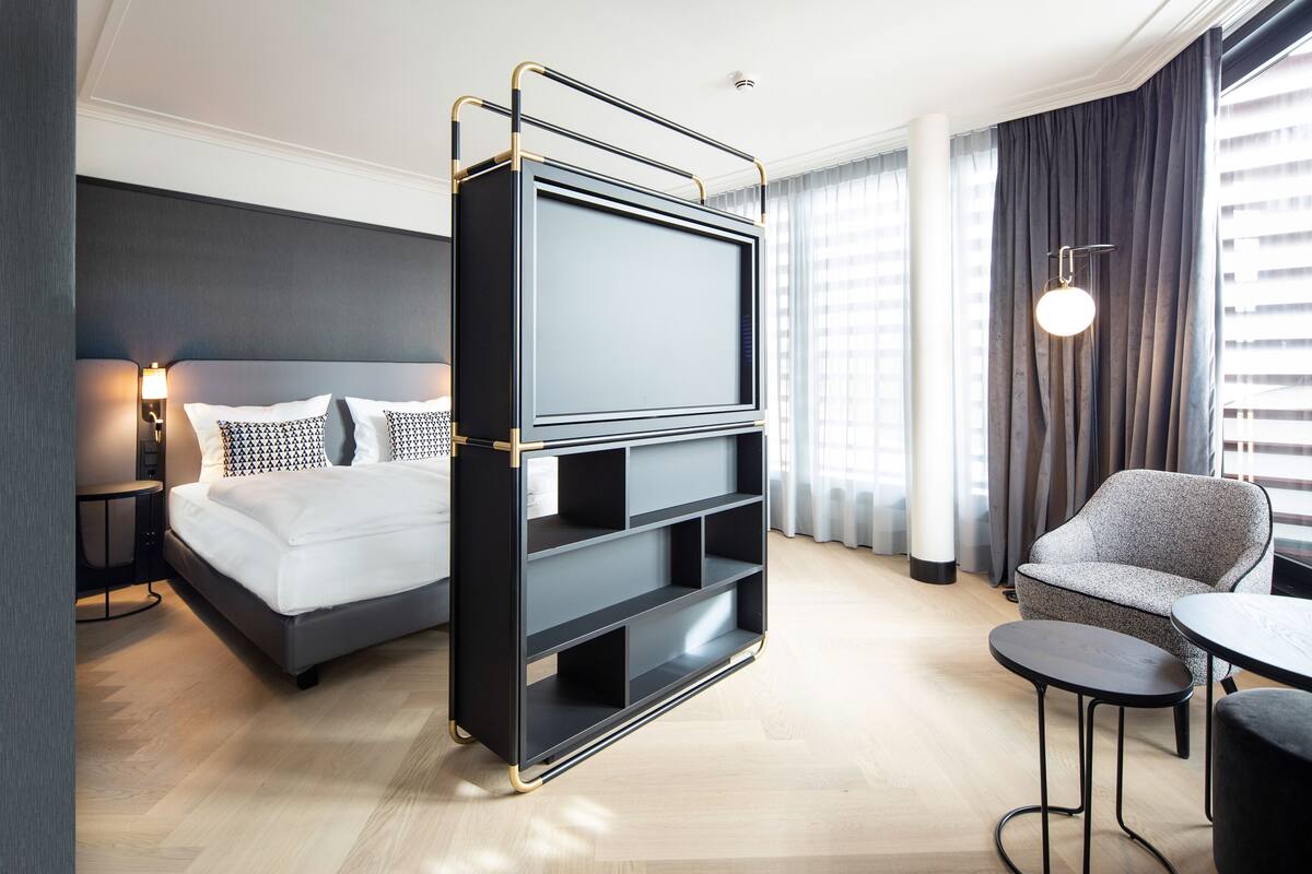 Quarto de hotel moderno com uma cama, um suporte para TV, uma cadeira e uma mesa pequena.