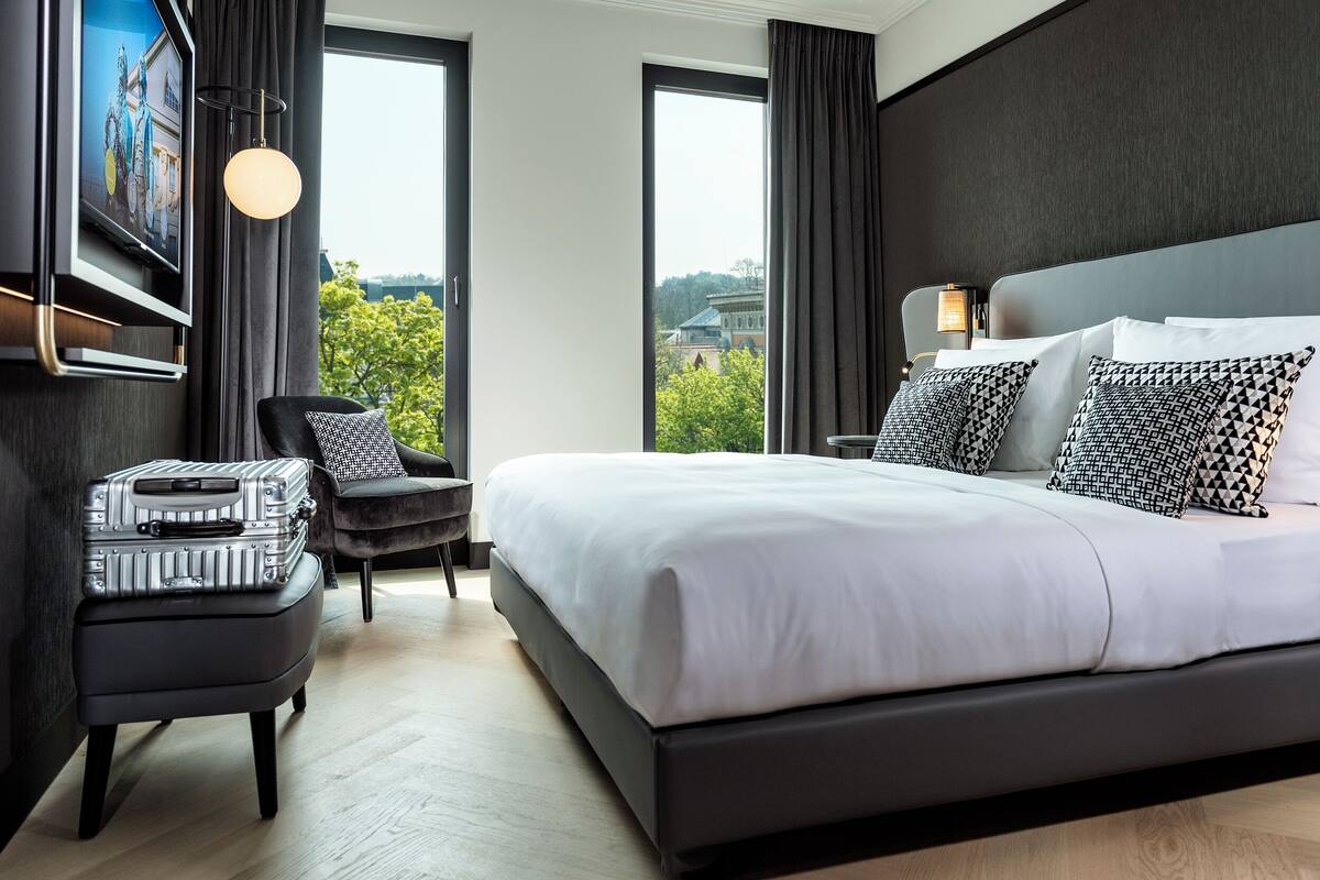 Quarto de hotel moderno com uma cama grande, televisão de tela plana na parede, pequeno espaço de estar com uma poltrona e um banquinho, e amplas janelas com vista para a cidade.