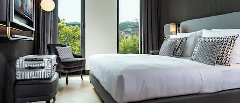 Une chambre d’hôtel moderne dotée d’un grand lit, d’un téléviseur à écran plat fixé au mur, d’un petit coin salon avec un fauteuil et un pouf, et de grandes fenêtres offrant une vue sur la ville.