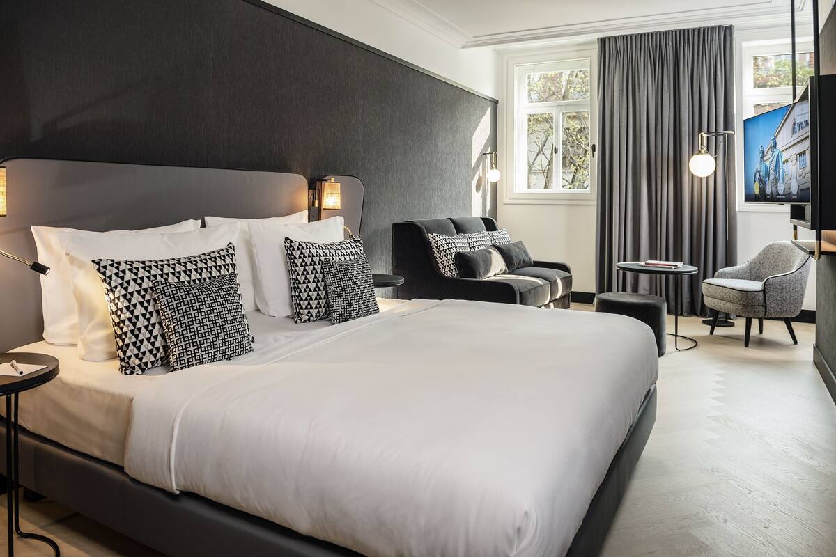 Quarto de hotel moderno com uma cama grande, cabeceira escura e um espaço de estar com uma cadeira e uma mesa pequena.