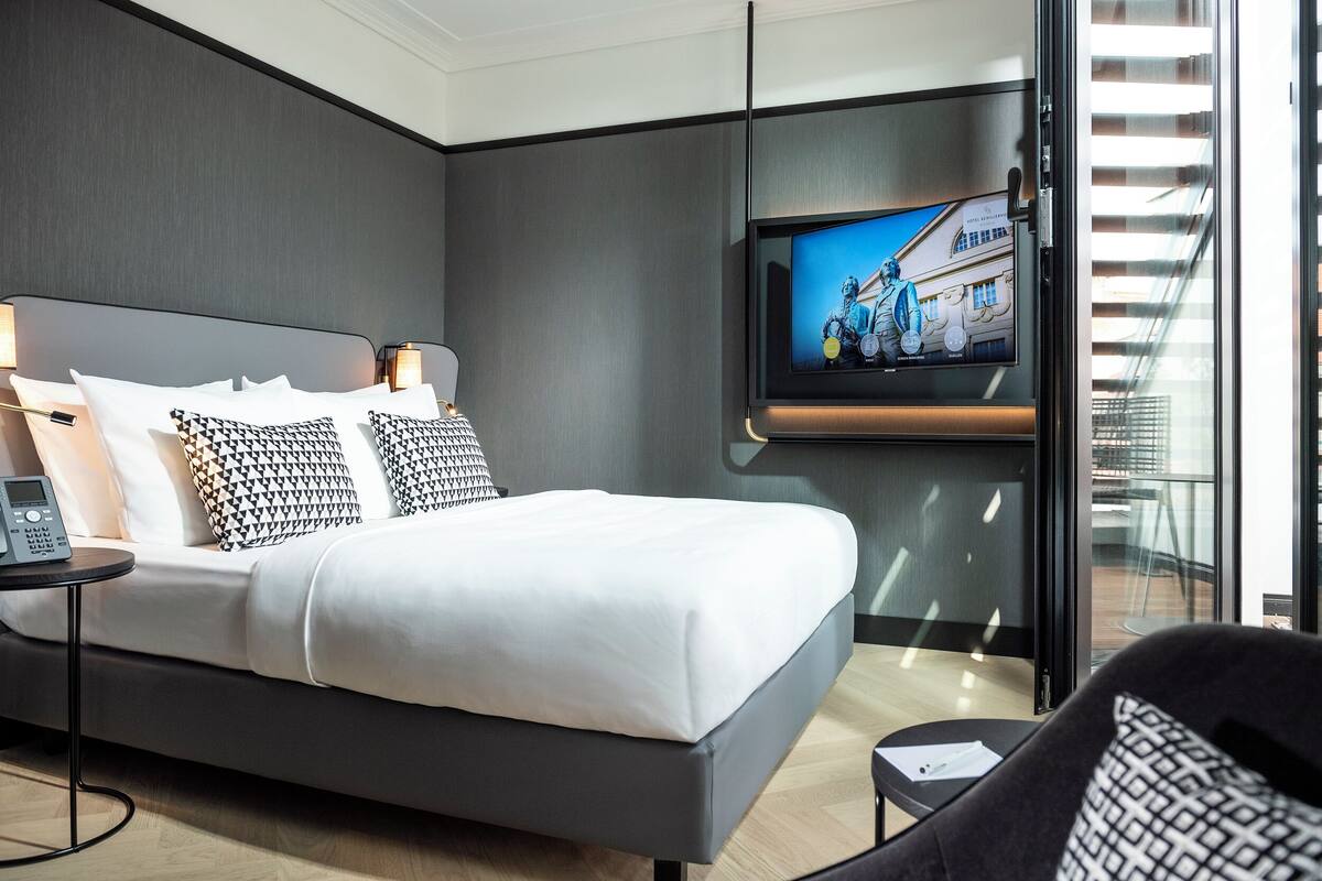Quarto de hotel moderno com uma cama grande, uma televisão presa na parede, uma mesa de cabeceira com telefone e vista para o exterior através de uma porta de vidro.