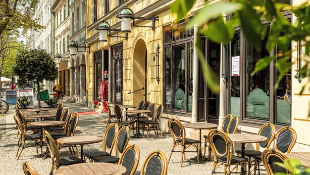 Un café al aire libre con sillas y mesas de mimbre, suelo adoquinado y un edificio con grandes ventanales y un letrero.