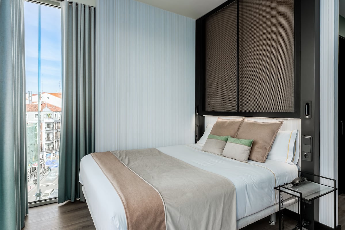 Ein modernes Hotelzimmer mit einem großen Bett, einem Nachttisch, einem Fenster mit Vorhängen und Blick auf die Stadtlandschaft.