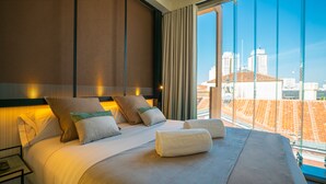 A hotel room with a large bed, two pillows, a nightstand, and a view of the city through large windows. - Akeah Hotel Gran Via (Madrid)