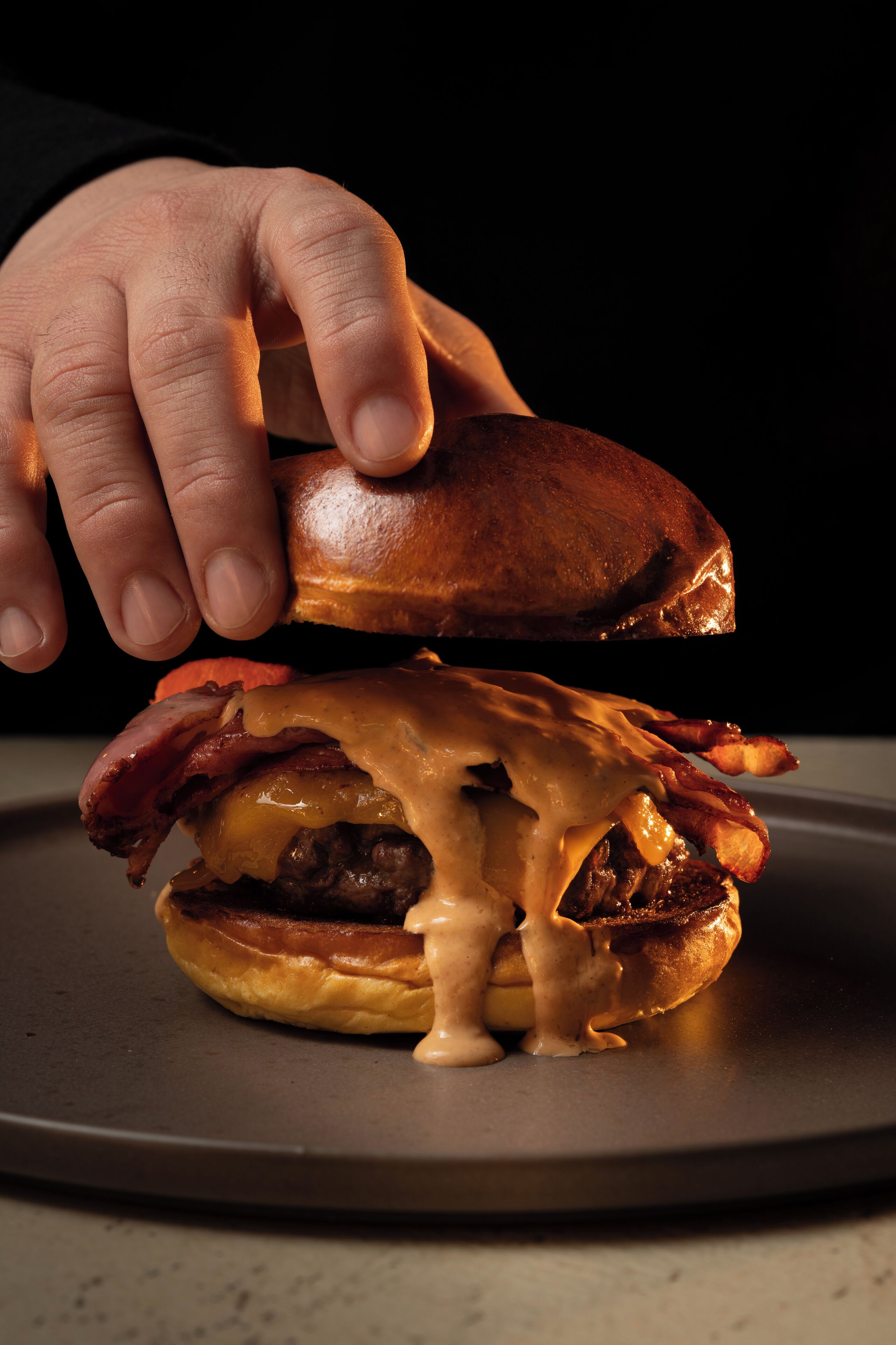 A hand holding a sandwich with melted cheese and bacon on a plate.