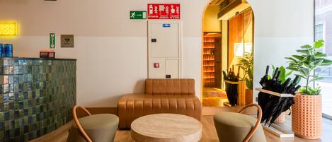 A modern lobby with a round table, two armchairs, a brown sofa, a reception desk, and an exit sign.