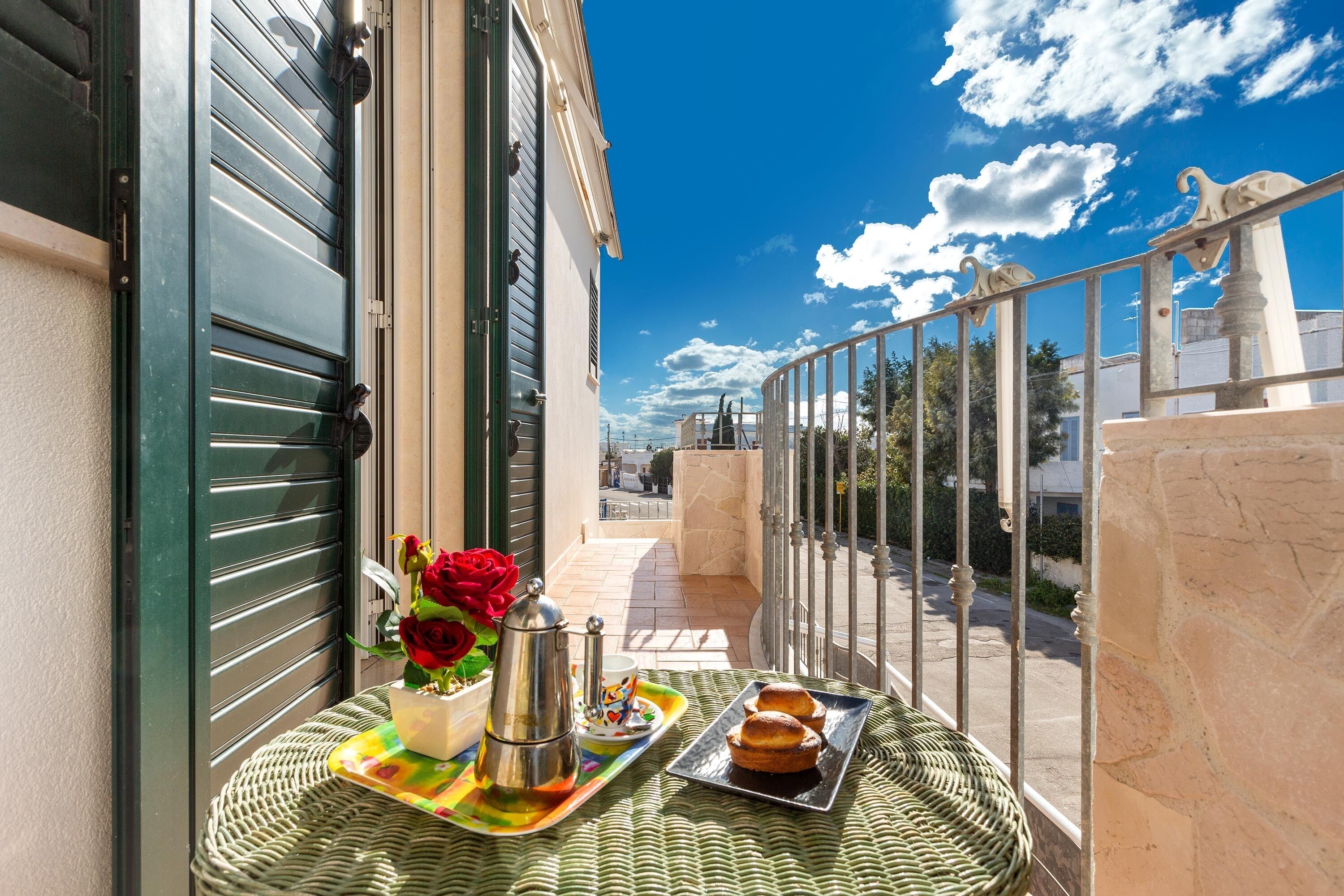 A balcony with a table set for breakfast, including pastries and a coffee pot, overlooking a street with buildings and a clear sky.