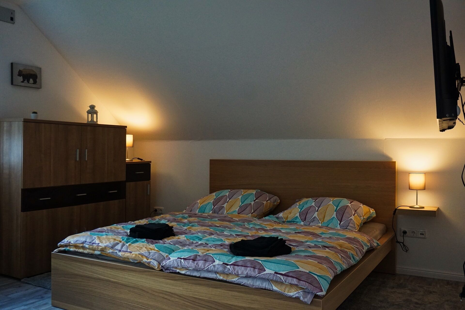 A bedroom with a wooden bed, two bedside lamps, a wardrobe, and a wall-mounted TV.