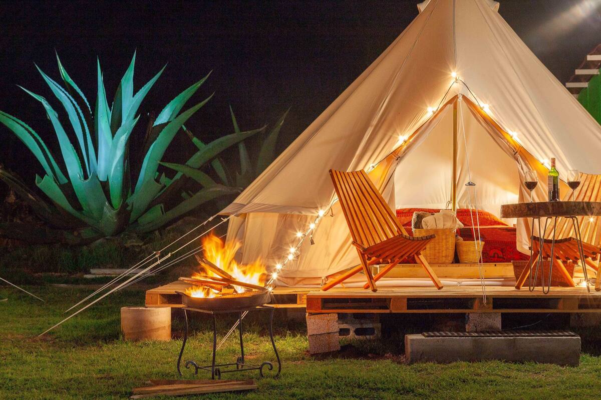 A tent with a fire pit and seating area, surrounded by greenery.