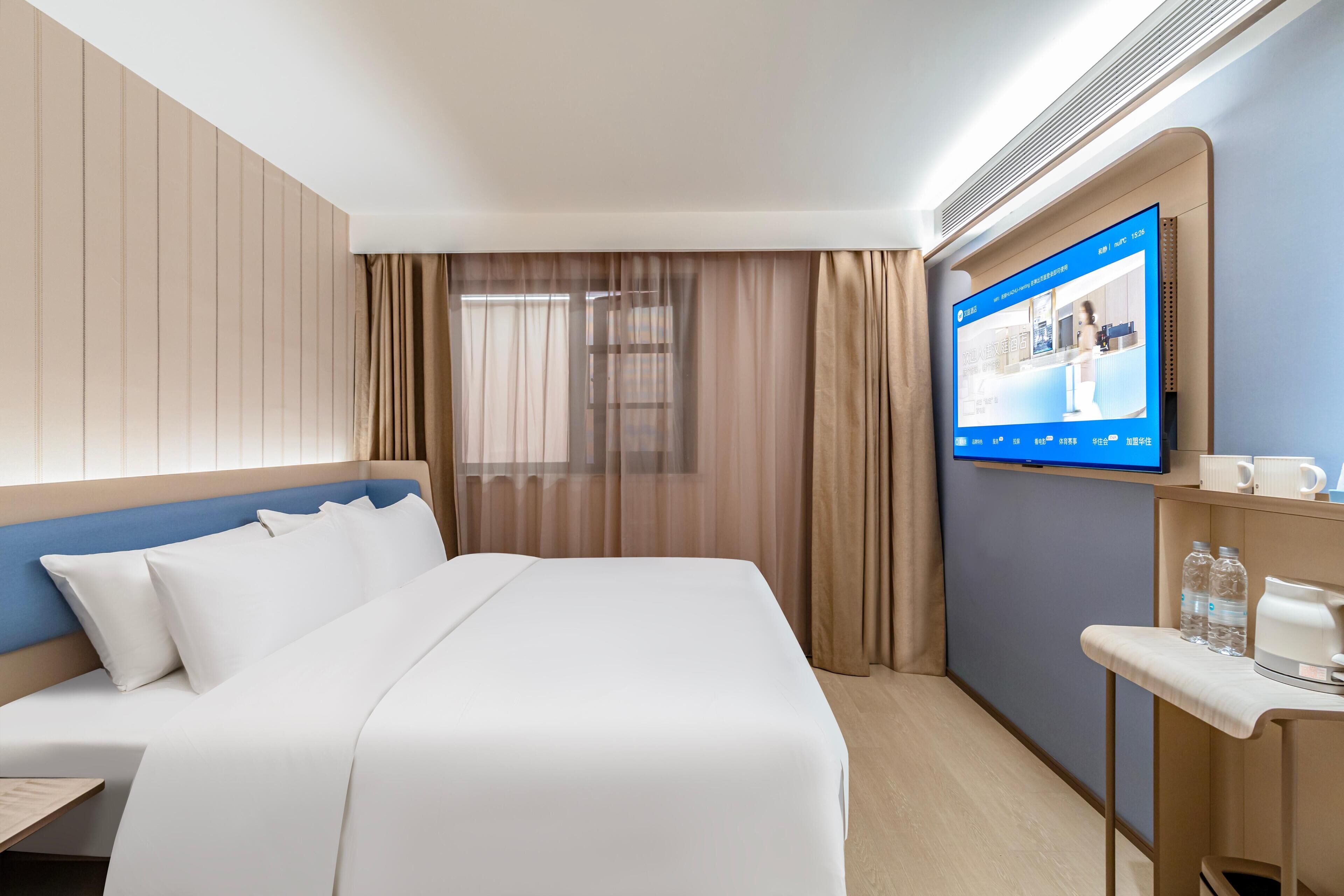 A hotel room with a bed, a TV mounted on the wall, a desk with a chair, and a window with curtains.