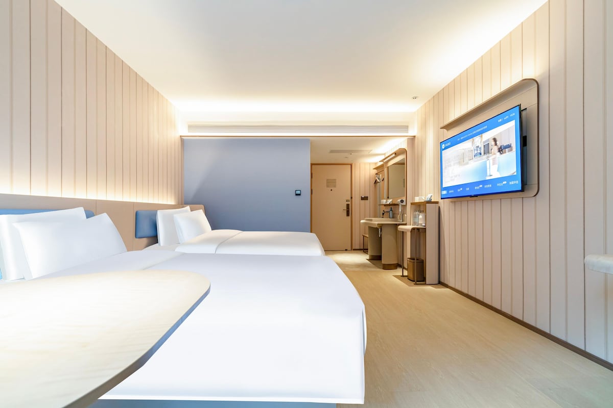 A hotel room with two beds, a television mounted on the wall, and a bathroom with a sink and mirror.