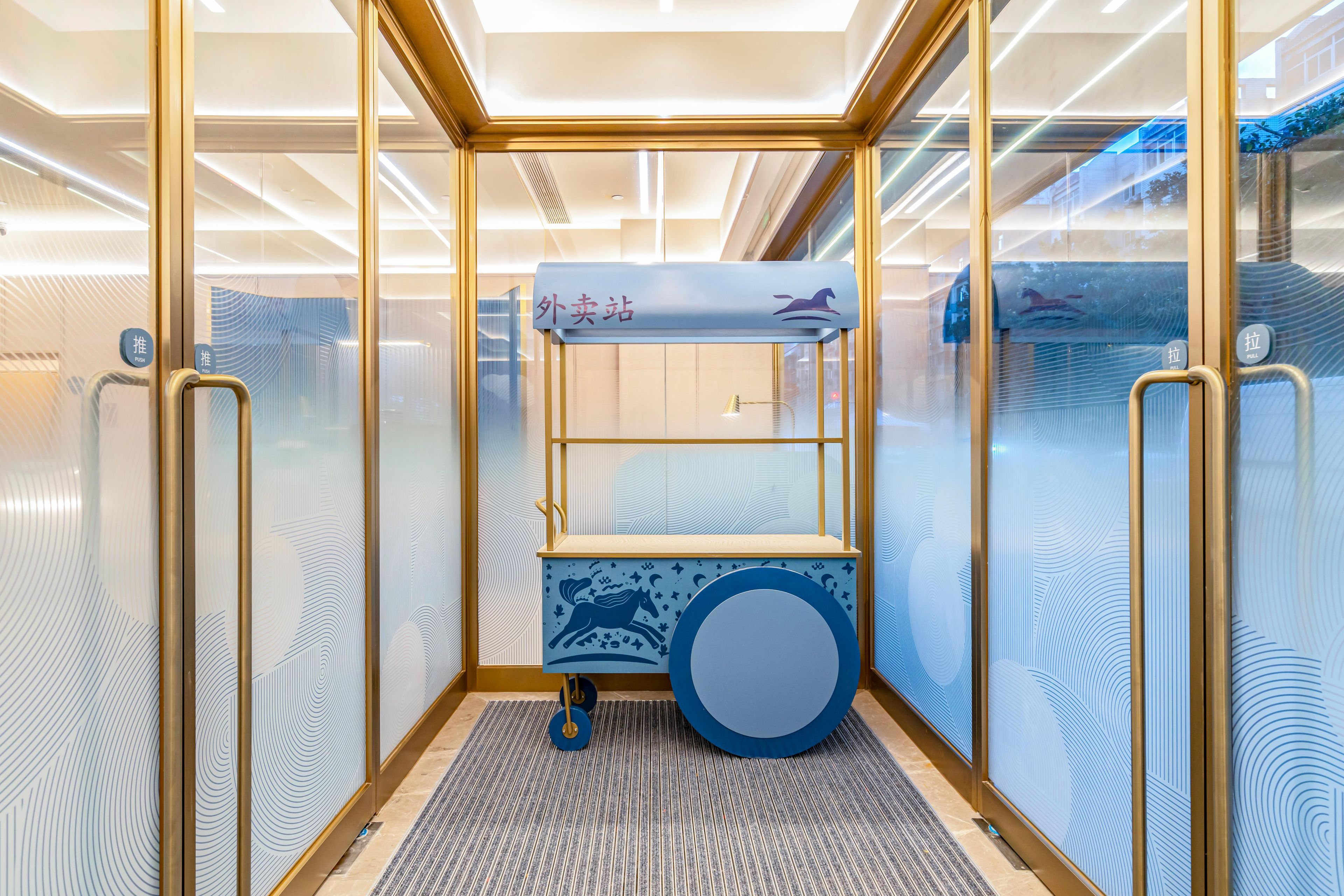 Un ascensor moderno con cabina de vidrio, piso de diseño azul y blanco, y un pequeño estante con una ilustración de un caballo.