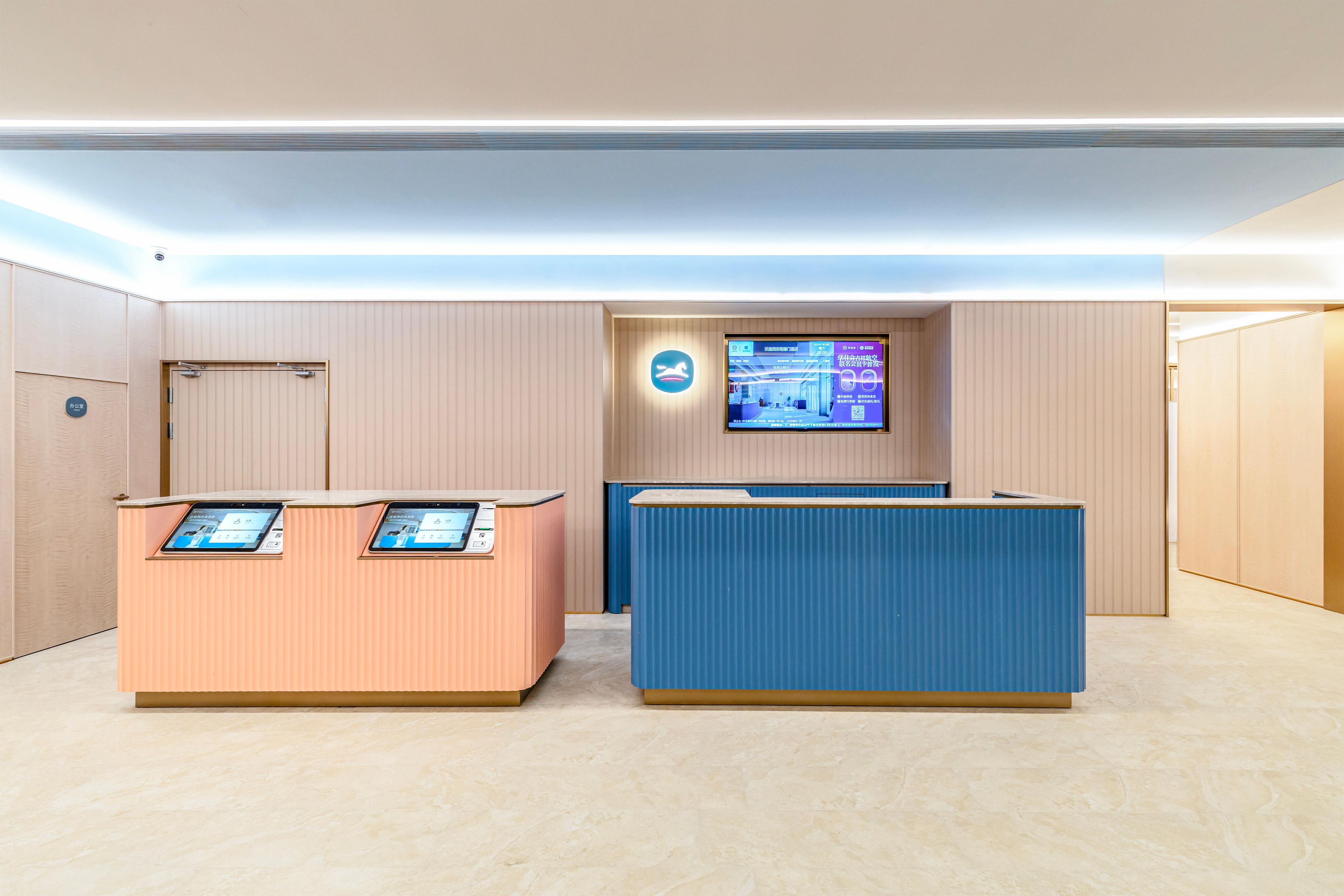 Un área de recepción con un quiosco de información digital, una pantalla de televisión y un mostrador azul.
