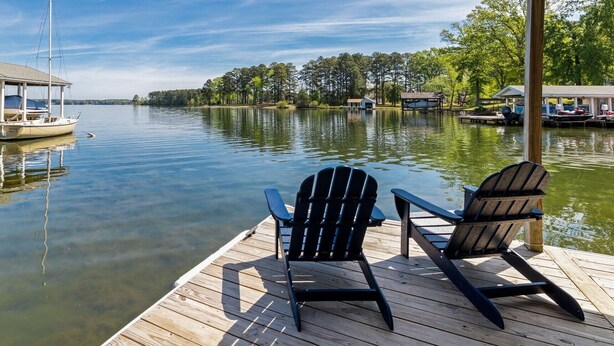 Une jetée en bois avec deux chaises Adirondack, donnant sur un lac paisible, entourée d’arbres et bordée de bateaux amarrés.
