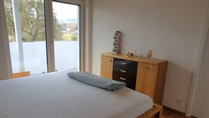 Un dormitorio con cama, cómoda de madera y ventana con vistas a casas y árboles.