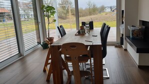 Un comedor con mesa y sillas de madera, ventanas amplias con persianas y vista a una zona residencial.
