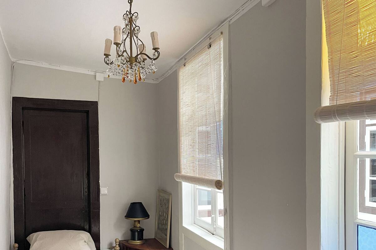 A bedroom with a bed, a wooden dresser, a nightstand with a lamp, a chandelier, and a window with blinds.