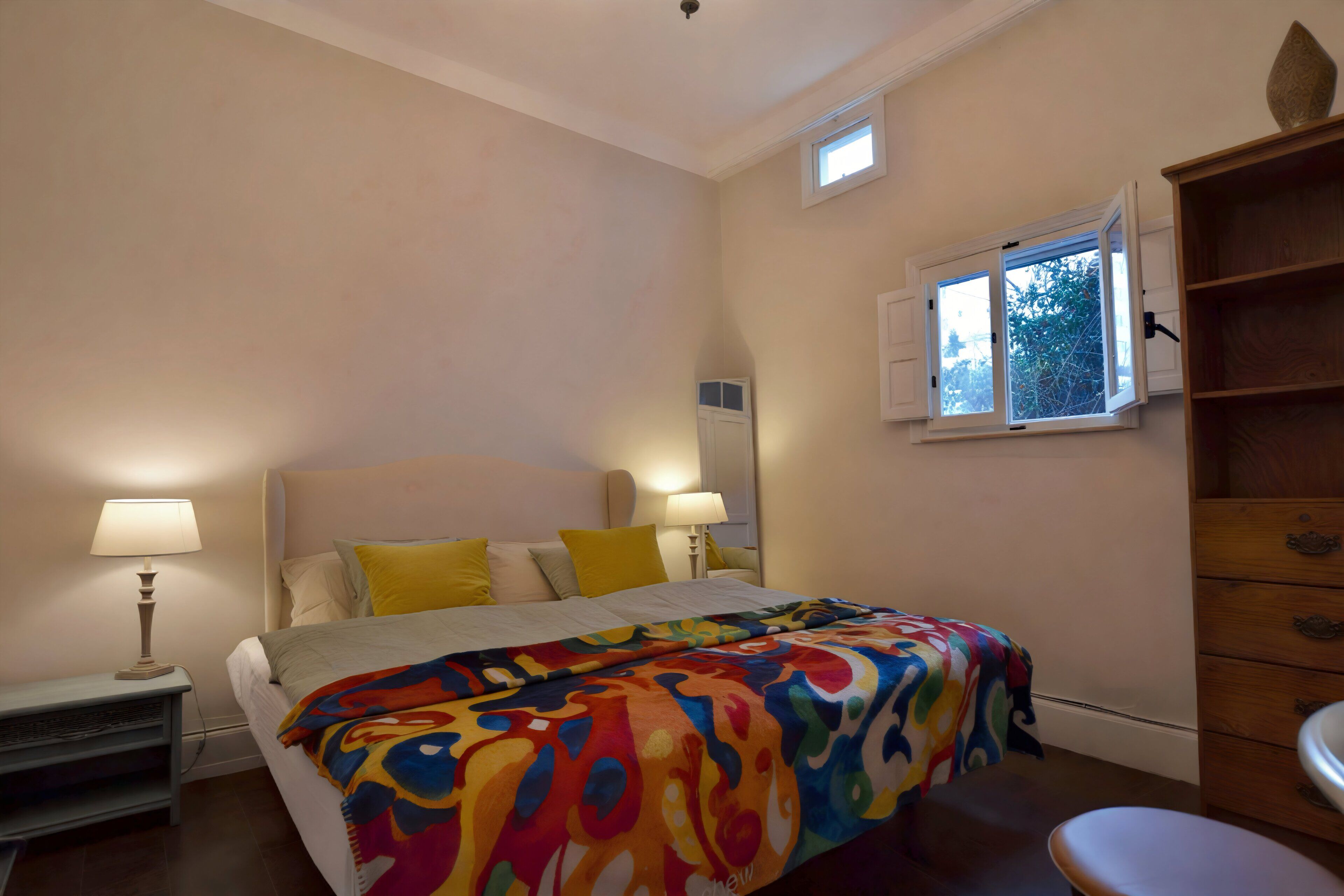 Quarto com uma cama, duas luminárias de cabeceira, uma janela com persianas e um aparador de madeira.