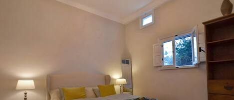 Quarto com uma cama, duas luminárias de cabeceira, uma janela com persianas e um aparador de madeira.