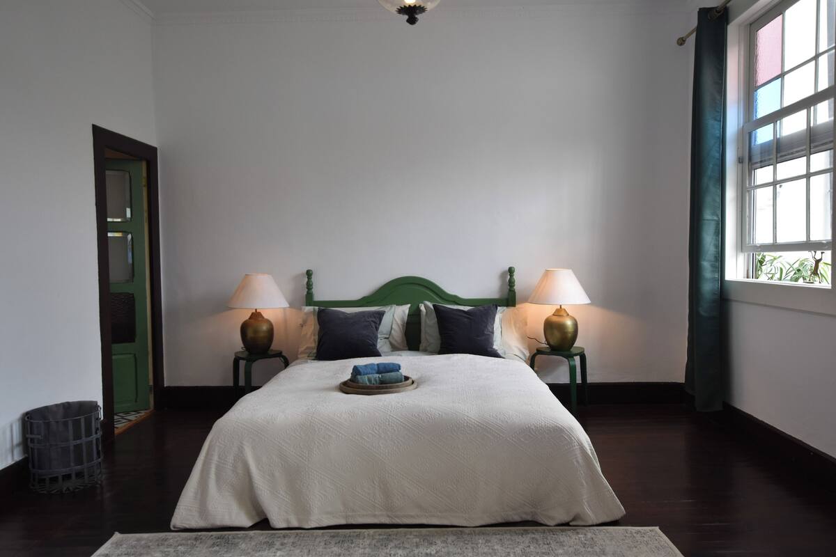 A bedroom with a large bed, two bedside lamps, a green headboard, and a window with curtains.