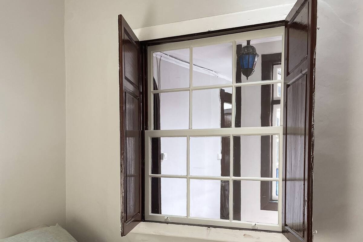 A bedroom with a bed, a window with open shutters, and a view of the outside.