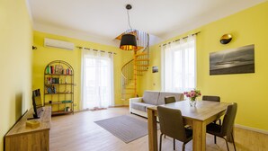 Ein hellgelbes Zimmer mit Esstisch, Stühlen, einer Couch, einer Wendeltreppe und einem Bücherregal.