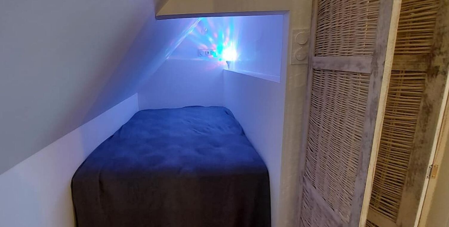 A small, narrow room with a single bed, a slanted ceiling, and a window with a light fixture.