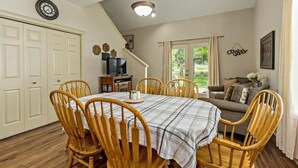 Un coin repas avec une table ronde, six chaises en bois, une horloge au mur et un escalier menant à un salon.