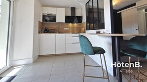 A modern kitchen with white cabinets, a black range hood, and a built-in oven. There is a bar area with a teal bar stool and a black chair. The kitchen has a large window providing natural light. - Town Hall Studio - Hyper-center Limoges (Limoges)