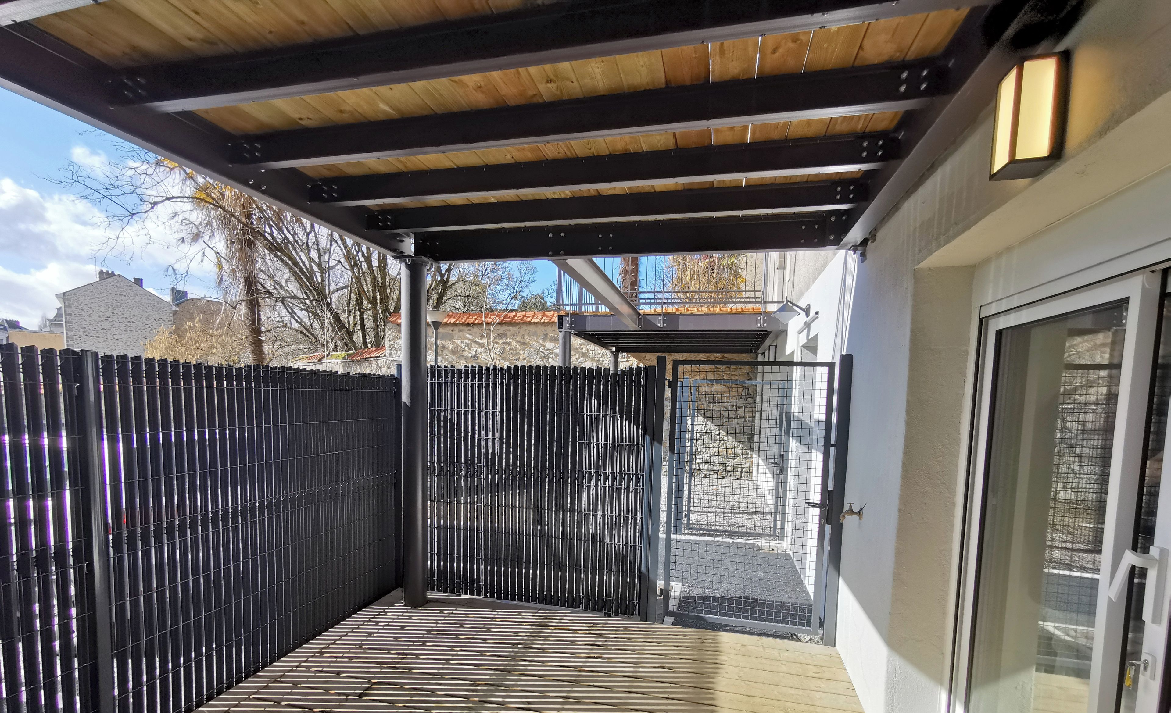 A covered patio area with a wooden deck, a metal fence, and a glass door leading to a building.