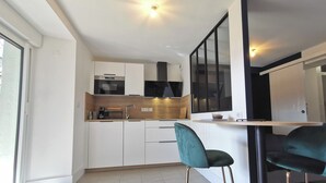 A modern kitchen with white cabinets, a black range hood, and a built-in oven. There is a bar area with a teal bar stool and a black chair. The kitchen has a large window providing natural light. - Town Hall Studio - Hyper-center Limoges (Limoges)