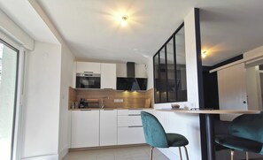 A modern kitchen with white cabinets, a black range hood, and a built-in oven. There is a bar area with a teal bar stool and a black chair. The kitchen has a large window providing natural light. - Town Hall Studio - Hyper-center Limoges (Limoges)