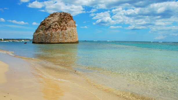 A clear beach with a large rock formation in the water.