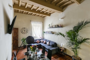 A living room with a dark grey sectional sofa, a dining table set with plates and glasses, a large potted plant, and a mounted flat-screen TV.