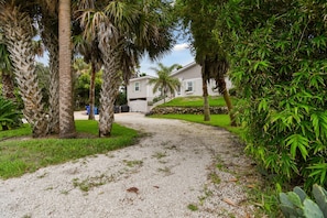 A gravel pathway leading to a residential building with a white exterior, surrounded by lush greenery including palm trees and bamboo.