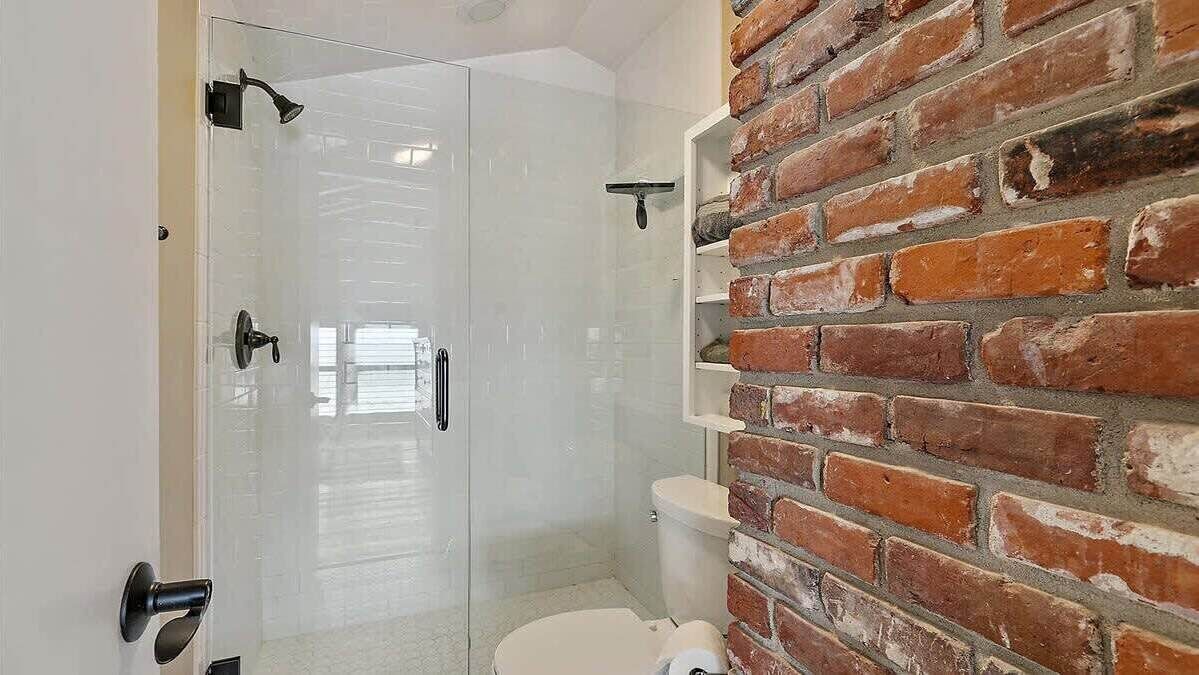 A bathroom with a glass shower enclosure, a white toilet, and a brick wall.