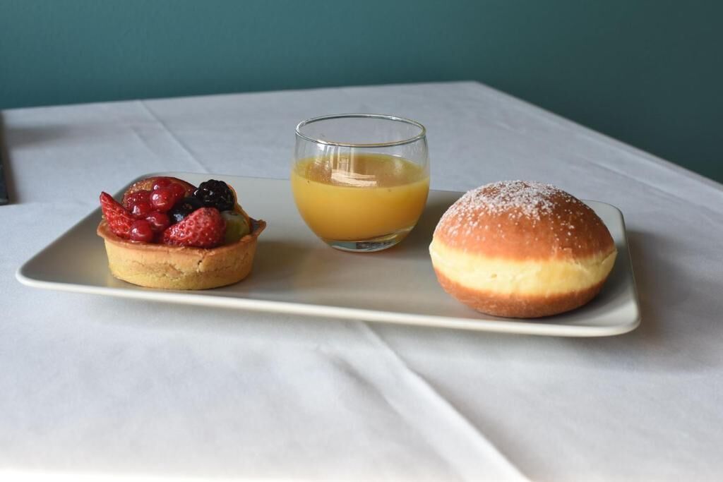 Une assiette à dessert avec une pâtisserie garnie de fruits, un verre de jus d’orange et un petit pain nature sur une nappe blanche.